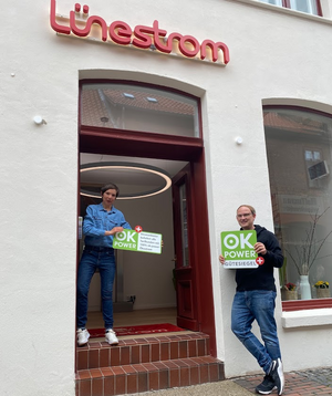 Jowana Lohmöller und Malte Steinmetz stehen im Eingang des Lünestrom ServiceCenters und halten jeweils ein ok-power+ Siegel.