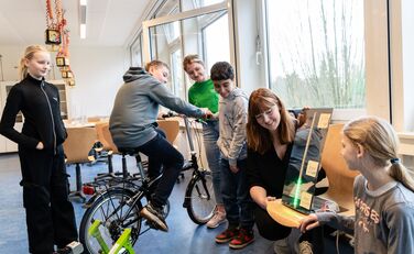 Ein Schüler sitzt auf einem Fahrrad, weitere Schüler*innen und Rebecca Baumer (SCHUBZ) sowie Lisa Persiel (Lünestrom) schauen auf einer Anzeige, wieviel Leistung erradelt wird.